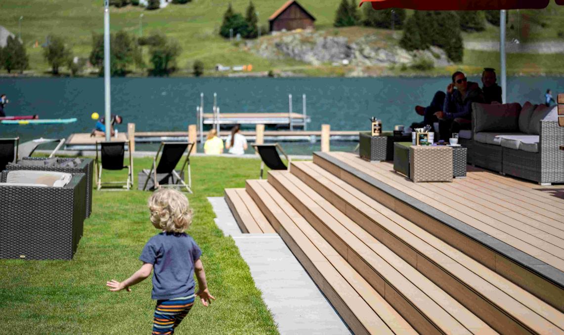 Enfant aux cheveux blonds courant sur une pelouse vers une terrasse en bois composite à plusieurs niveaux, avec mobilier de jardin en résine tressée, parasol rouge, et un lac bleu avec ponton en arrière-plan